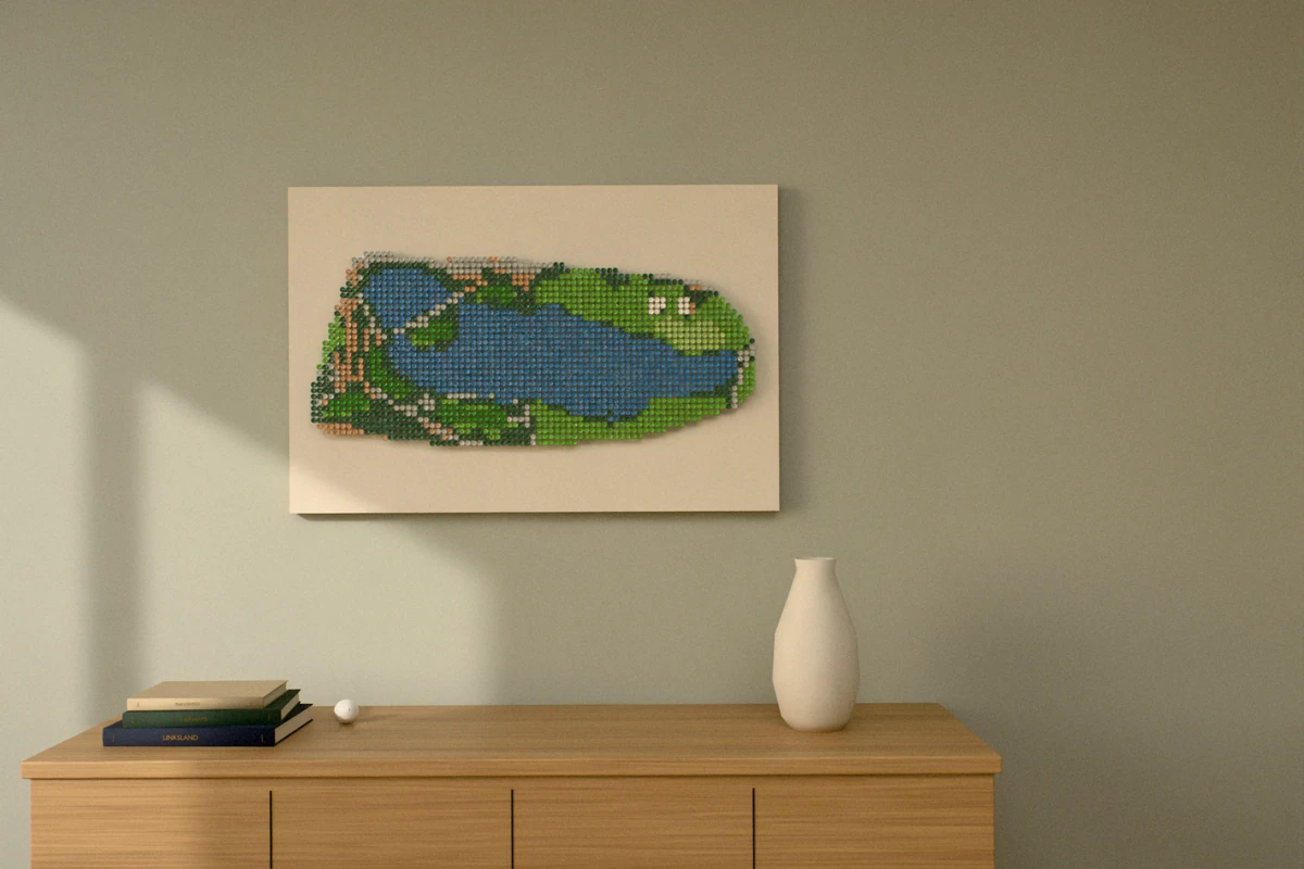 A golf-tee picture of a par 3 played over a long lake, hung wide on a sage wall above a wooden sideboard with books and a vase.